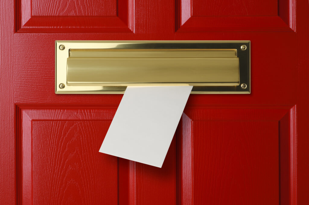 A white envelope coming through a brass letterbox on a red front door